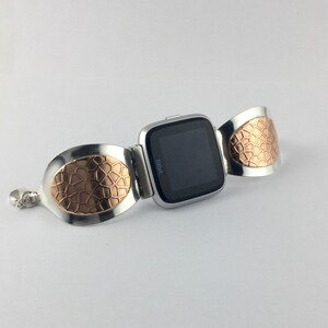 May include: A silver and rose gold Fitbit watch band with a textured design. The band is made of two oval-shaped pieces that wrap around the wrist and connect to the watch face.