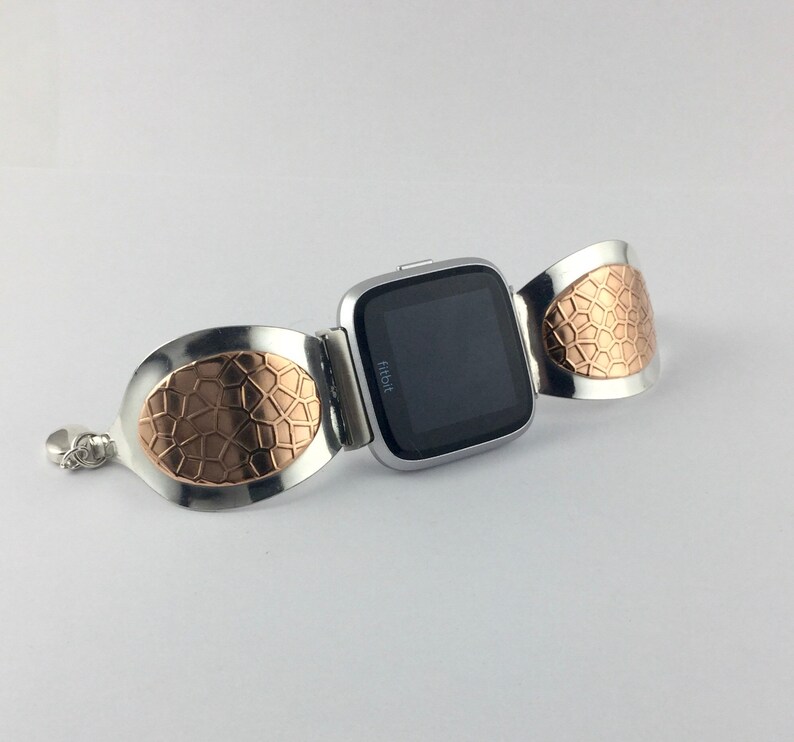 May include: A silver and rose gold Fitbit watch band with a textured design. The band is made of two oval-shaped pieces that wrap around the wrist and connect to the watch face.