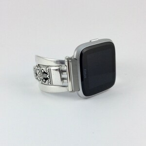 May include: Silver metal watch band with an intricate floral design. The band is attached to a silver Fitbit smartwatch with a black screen.
