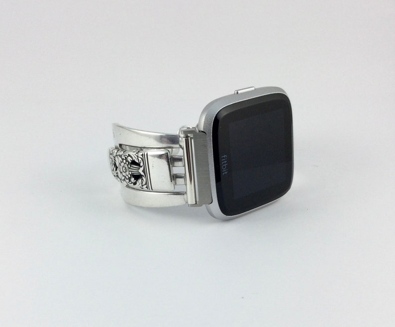May include: Silver metal watch band with an intricate floral design. The band is attached to a silver Fitbit smartwatch with a black screen.
