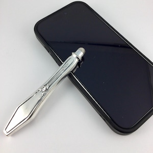 May include: A silver-toned stylus with an ornate, vintage-style handle featuring embossed detailing and a rounded tip, positioned diagonally across the black screen of a rectangular smartphone with a black protective case.