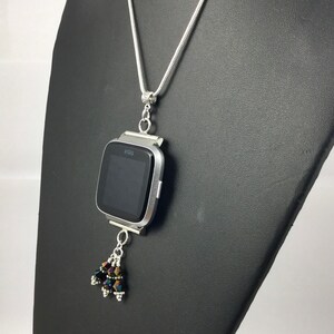 Puede incluir: Collar plateado con un colgante de reloj inteligente Fitbit Versa. El reloj tiene una pantalla negra y est&aacute; adornado con un grupo de cuentas iridiscentes. El collar se exhibe en un maniqu&iacute; negro.