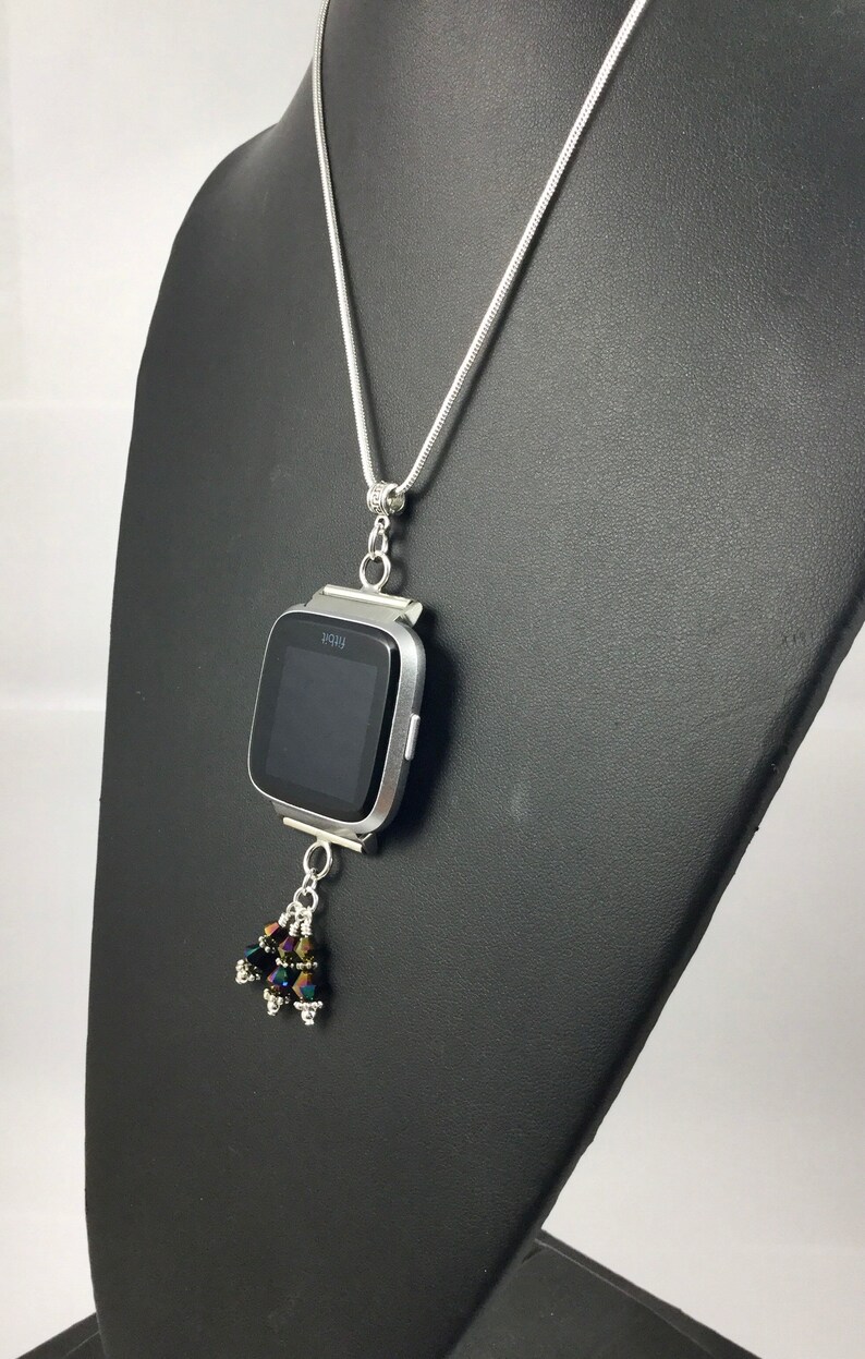 Puede incluir: Collar plateado con un colgante de reloj inteligente Fitbit Versa. El reloj tiene una pantalla negra y est&aacute; adornado con un grupo de cuentas iridiscentes. El collar se exhibe en un maniqu&iacute; negro.