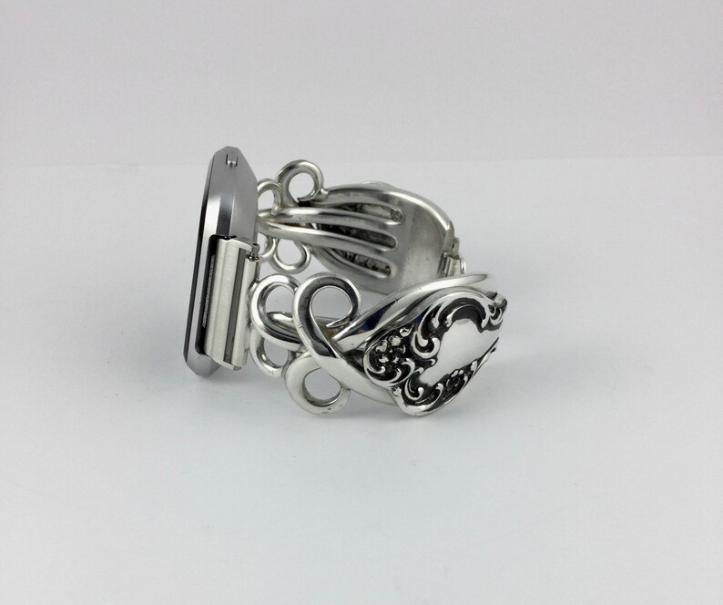 May include: Silver bracelet made from a vintage silverware fork. The bracelet is designed to fit a watch or smart watch. The bracelet has a decorative design with a floral pattern.