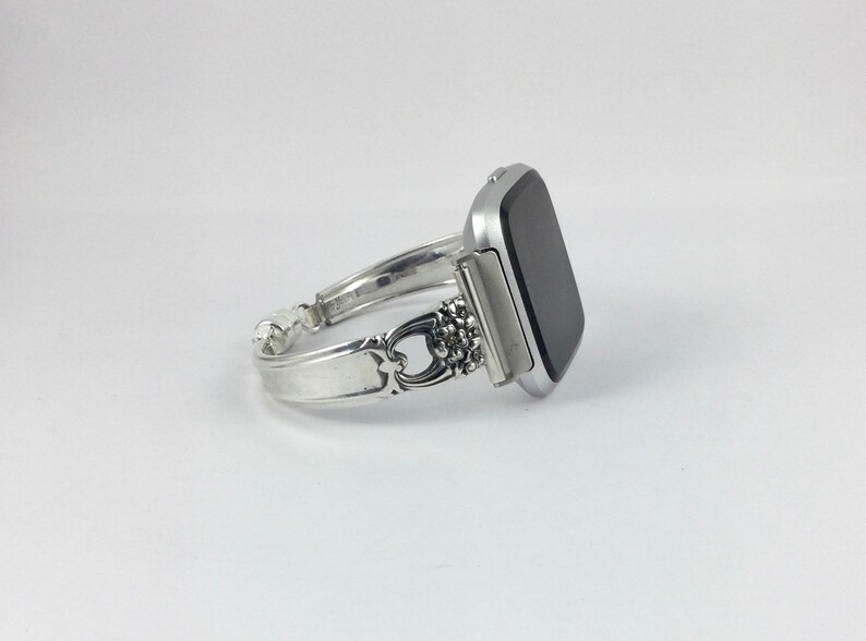 May include: A silver watch band made from a repurposed silverware piece. The band is attached to a silver smartwatch with a black screen.
