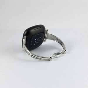 Puede incluir: Un brazalete plateado con un dise&ntilde;o intrincado, con un reloj inteligente negro adjunto.
