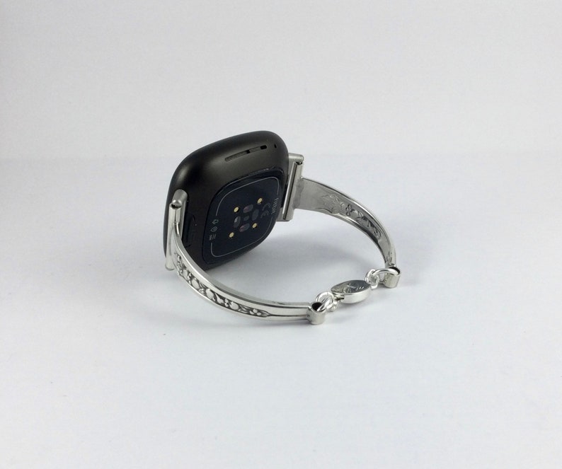 Puede incluir: Un brazalete plateado con un dise&ntilde;o intrincado, con un reloj inteligente negro adjunto.