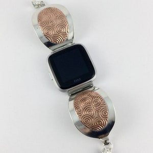 May include: A silver and rose gold Fitbit watch band with a swirl design. The band is made of two oval-shaped pieces that connect to the watch face.