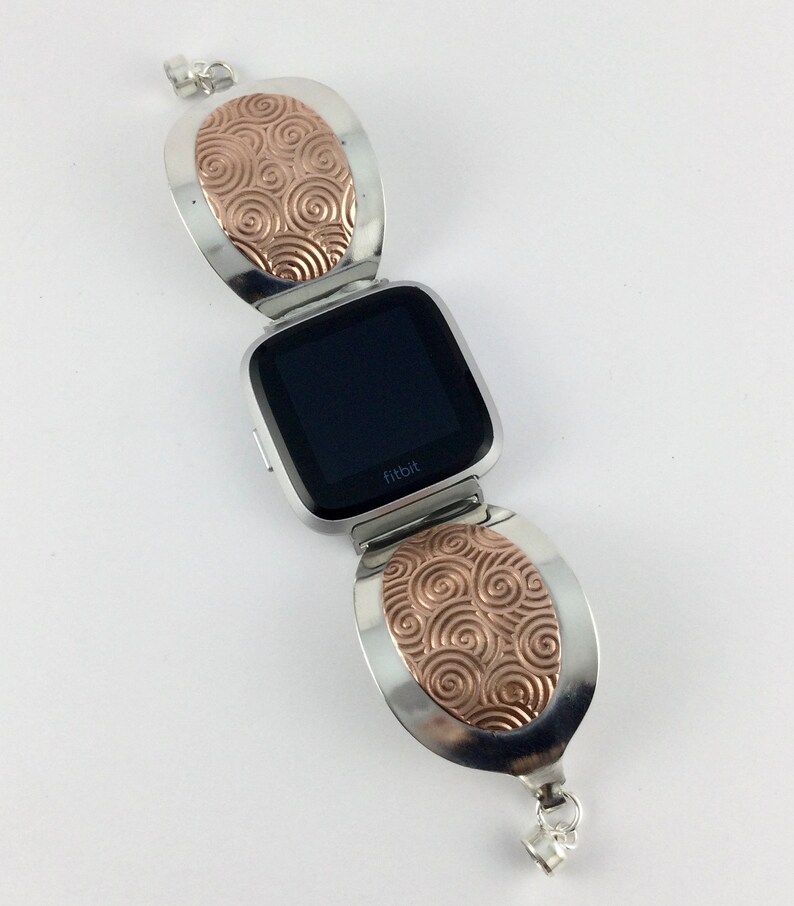 May include: A silver and rose gold Fitbit watch band with a swirl design. The band is made of two oval-shaped pieces that connect to the watch face.