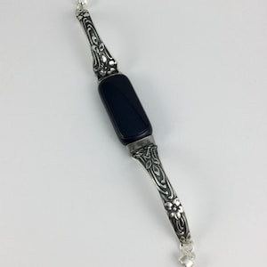 May include: A fitness tracker with a rectangular black screen and silver-toned metal bands featuring an embossed floral and leaf pattern. The metallic segments are connected to the central display unit, creating a decorative bracelet-style accessory.