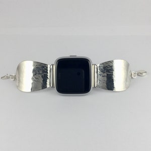 May include: Silver watch band with a hammered metal design. The band is attached to a square-shaped smartwatch with a black screen.