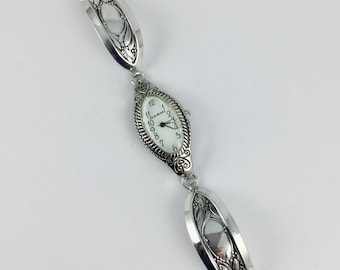 Könnte beinhalten: Silberfarbene Uhr mit ovalem Zifferblatt und weißem Zifferblatt mit schwarzen Ziffern und Zeigern. Die Uhr ist mit zwei dekorativen silbernen Bändern mit floralen Designs verbunden. Auf dem Zifferblatt steht "Narm".