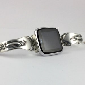 May include: A silver Fitbit smartwatch with a custom-made band crafted from a vintage silverware spoon. The band features intricate floral designs and a polished oval shape.