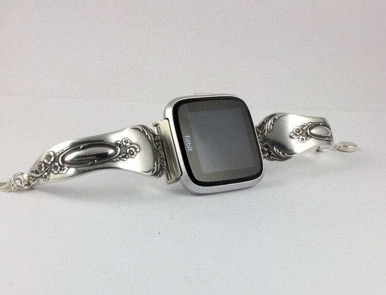 May include: A silver Fitbit smartwatch with a custom-made band crafted from a vintage silverware spoon. The band features intricate floral designs and a polished oval shape.