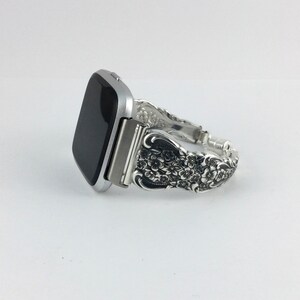 May include: Silver-toned smartwatch with a decorative band. The band is made from ornate silverware, featuring floral and scroll designs. The watch face is black, and the band is approximately 7 inches in length.