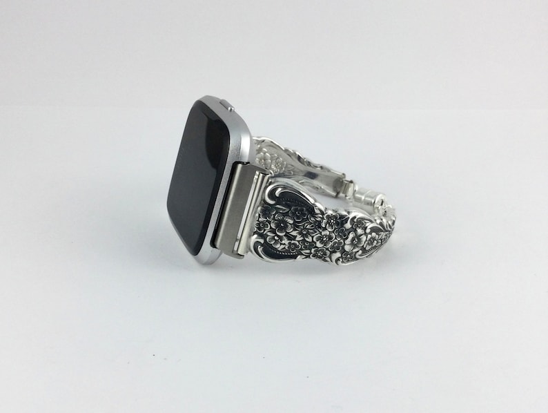 May include: Silver-toned smartwatch with a decorative band. The band is made from ornate silverware, featuring floral and scroll designs. The watch face is black, and the band is approximately 7 inches in length.