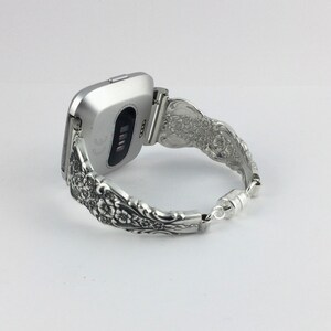 May include: Silver-toned Fitbit watch with a decorative band. The band is made from ornate silverware, featuring floral patterns. The watch is connected to the band with a hinge, and the band closes with a magnetic clasp.