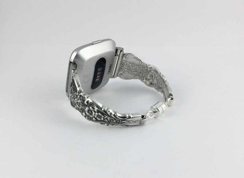 May include: Silver-toned Fitbit watch with a decorative band. The band is made from ornate silverware, featuring floral patterns. The watch is connected to the band with a hinge, and the band closes with a magnetic clasp.