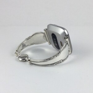 May include: A silver smartwatch with a silver spoon bracelet. The watch face is not visible, but the back of the watch is visible, showing the heart rate sensor.