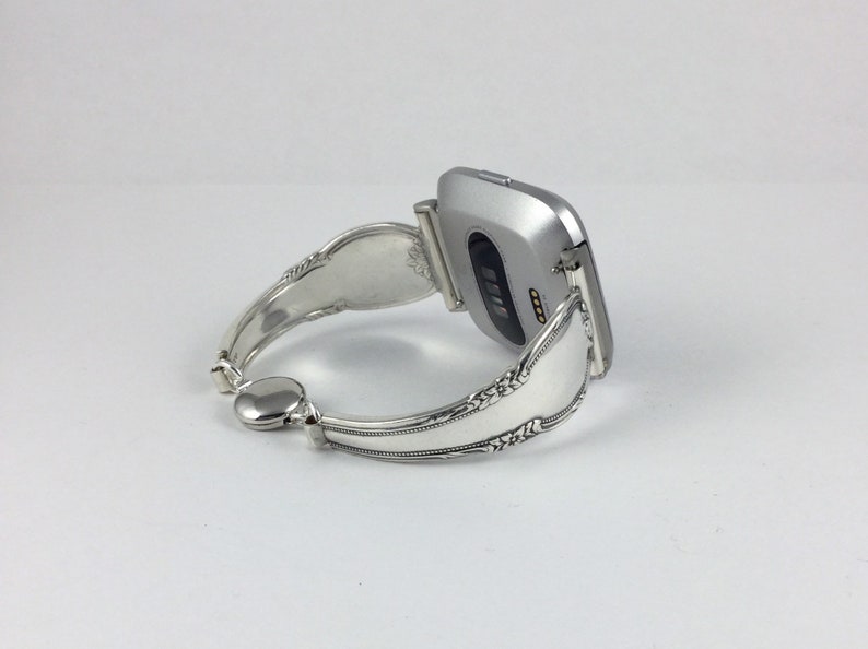 May include: A silver smartwatch with a silver spoon bracelet. The watch face is not visible, but the back of the watch is visible, showing the heart rate sensor.