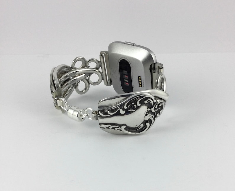 May include: A silver smartwatch with a silver spoon band. The watch face is black with a white digital display. The spoon band is intricately designed with a floral pattern.
