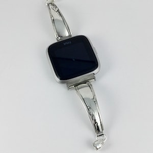 Peut inclure: Un bracelet de montre argent&eacute; fabriqu&eacute; &agrave; partir d'une pi&egrave;ce de couverts r&eacute;utilis&eacute;e. Le bracelet est con&ccedil;u pour s'adapter &agrave; une montre connect&eacute;e de forme carr&eacute;e. Le bracelet pr&eacute;sente un design orn&eacute; avec un motif floral.