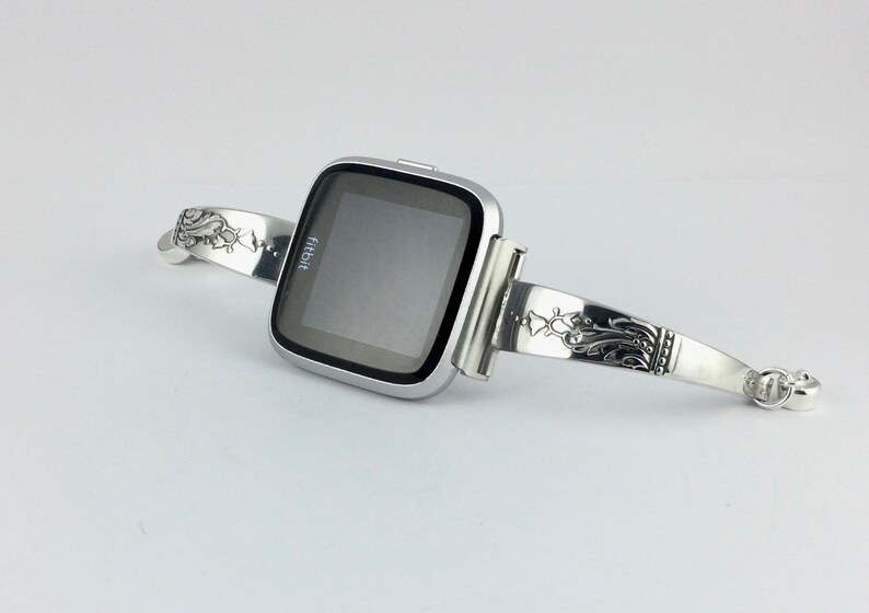 Op de afbeelding: Een zilveren Fitbit smartwatch met een zwart scherm is bevestigd aan een zilveren armband met een sierlijk ontwerp.