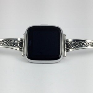 Puede incluir: Pulsera de reloj plateada con dise&ntilde;o floral. La pulsera est&aacute; dise&ntilde;ada para adaptarse a un reloj inteligente de forma cuadrada. La esfera del reloj es negra y muestra la palabra "fitbit".