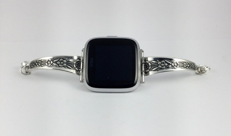 Puede incluir: Pulsera de reloj plateada con dise&ntilde;o floral. La pulsera est&aacute; dise&ntilde;ada para adaptarse a un reloj inteligente de forma cuadrada. La esfera del reloj es negra y muestra la palabra "fitbit".