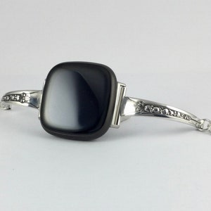 Puede incluir: Una pulsera de plata con una piedra de &aacute;gata negra y blanca. La pulsera tiene un dise&ntilde;o decorativo en la banda.