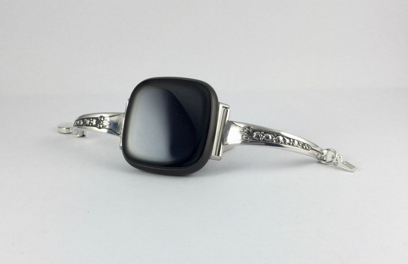Puede incluir: Una pulsera de plata con una piedra de &aacute;gata negra y blanca. La pulsera tiene un dise&ntilde;o decorativo en la banda.