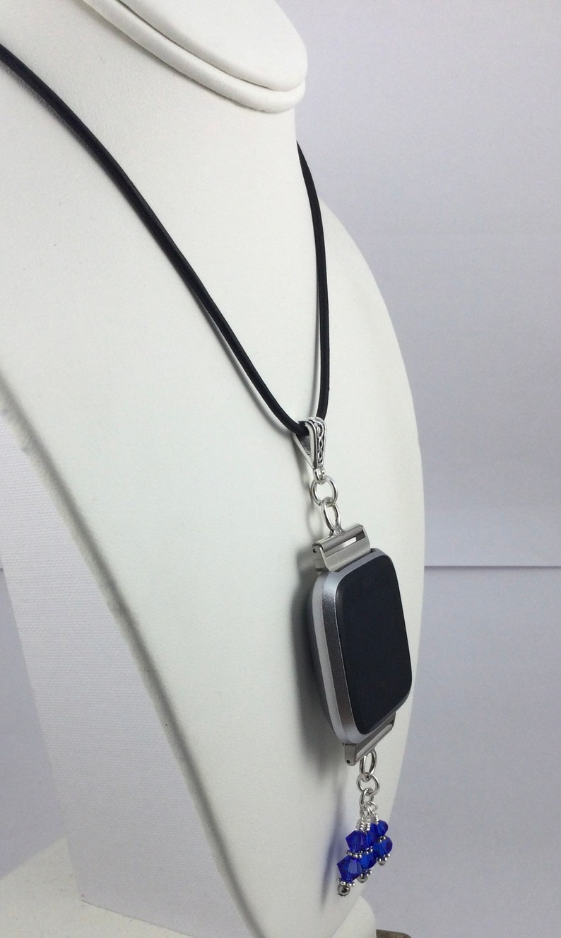 May include: A silver-toned smartwatch pendant necklace with a black screen, suspended from a black cord. The pendant features a cluster of blue crystal beads. The necklace is displayed on a white mannequin.