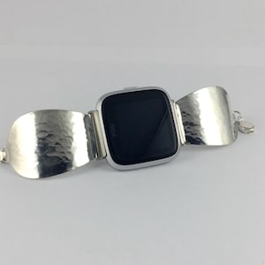 May include: A silver Fitbit smartwatch with a black screen is attached to a silver hammered metal bracelet.