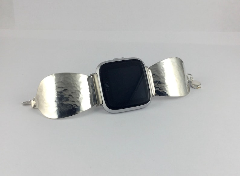 May include: A silver Fitbit smartwatch with a black screen is attached to a silver hammered metal bracelet.