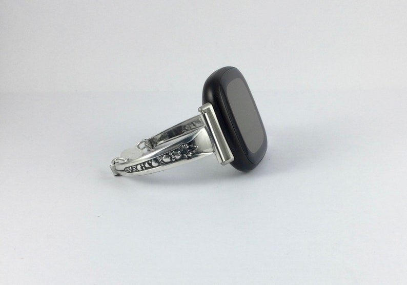 Puede incluir: Una pulsera de reloj plateada con un intrincado dise&ntilde;o floral. La pulsera est&aacute; unida a un reloj inteligente negro con una pantalla gris oscura.