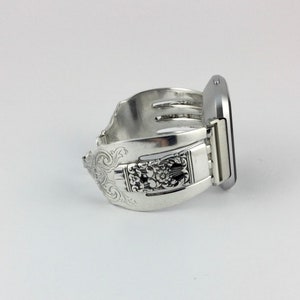 May include: A silver watch band made from a repurposed fork with an ornate design. The band is attached to a silver smartwatch.