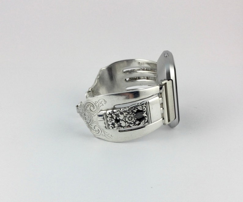 May include: A silver watch band made from a repurposed fork with an ornate design. The band is attached to a silver smartwatch.
