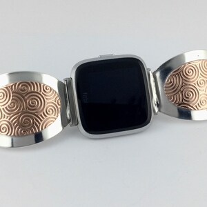 May include: A silver and rose gold watch band with a swirling pattern. The band is designed to fit a square-shaped smartwatch.