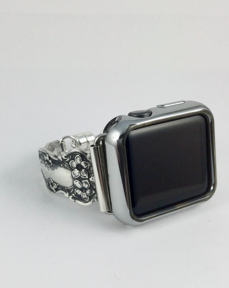 May include: A silver-toned smartwatch with a black screen and a metallic watch band featuring an embossed floral pattern. The band is constructed from a repurposed silver-colored spoon handle with intricate flower details and a polished finish.