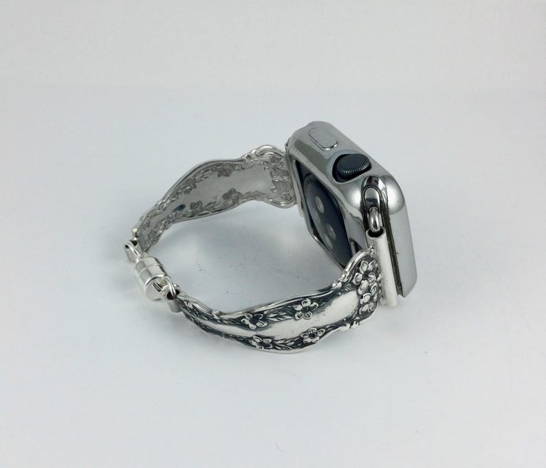 May include: A silver-toned smartwatch with a metallic finish and a black circular sensor on the back. The watch is attached to a decorative silver-colored metal band featuring an embossed floral pattern and a magnetic clasp closure mechanism.