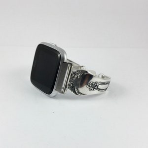 May include: Silver-toned Fitbit smartwatch with a unique, ornate, silver-toned band made from a repurposed spoon.