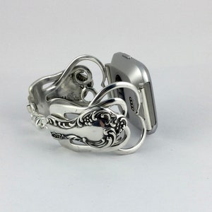 Puede incluir: Una pulsera de reloj inteligente plateada hecha de cubiertos reutilizados. La pulsera tiene forma de dos corazones y tiene un dise&ntilde;o decorativo. La pulsera est&aacute; unida a un reloj inteligente plateado.