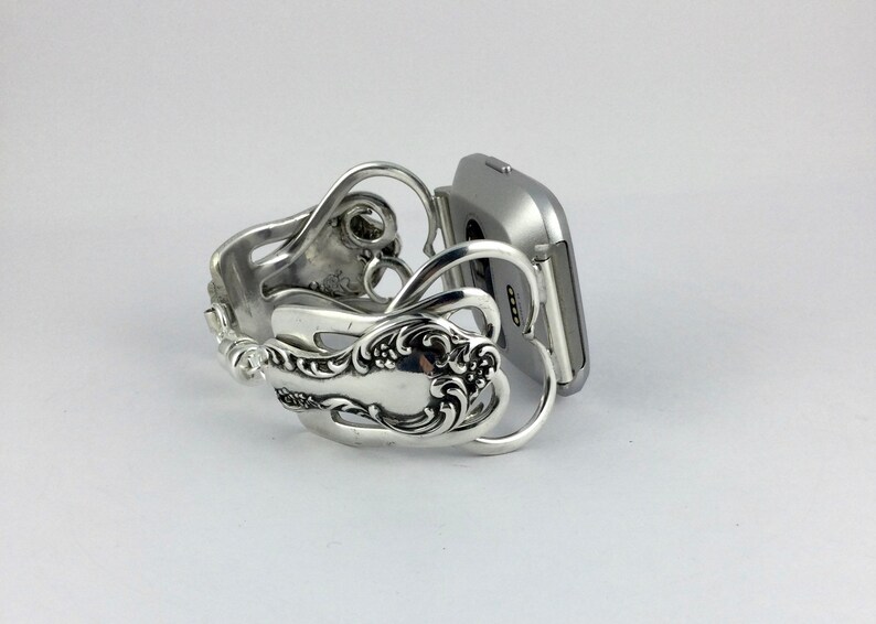 Puede incluir: Una pulsera de reloj inteligente plateada hecha de cubiertos reutilizados. La pulsera tiene forma de dos corazones y tiene un dise&ntilde;o decorativo. La pulsera est&aacute; unida a un reloj inteligente plateado.