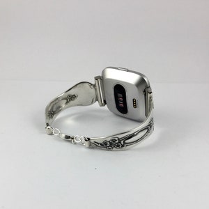 May include: Silver watch band made from a repurposed spoon with floral details. The band is attached to a silver smartwatch with a black screen.