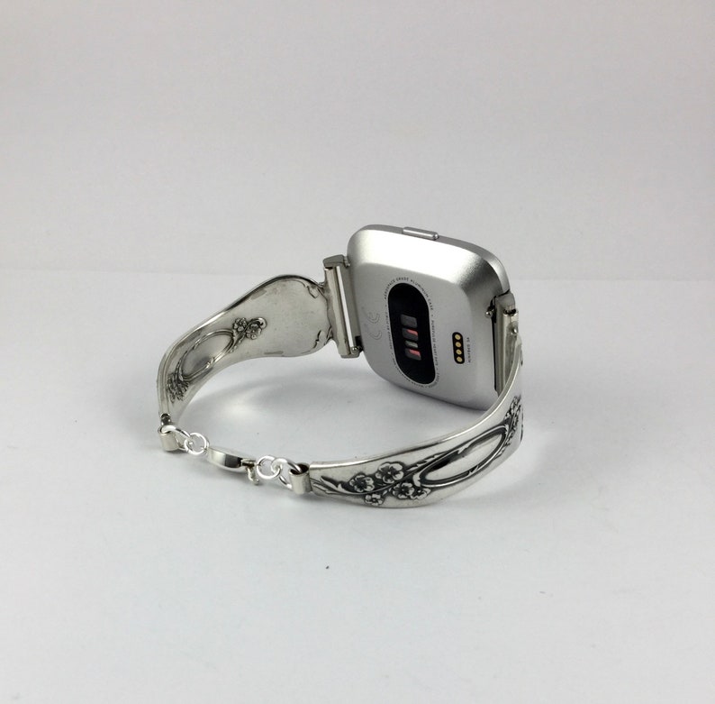 May include: Silver watch band made from a repurposed spoon with floral details. The band is attached to a silver smartwatch with a black screen.
