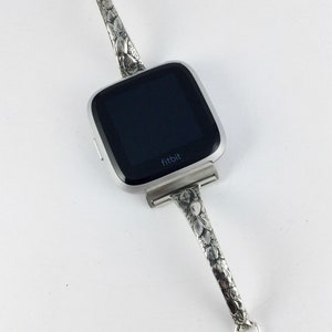 May include: A silver Fitbit smartwatch with a floral patterned band. The band has a decorative charm on each end.