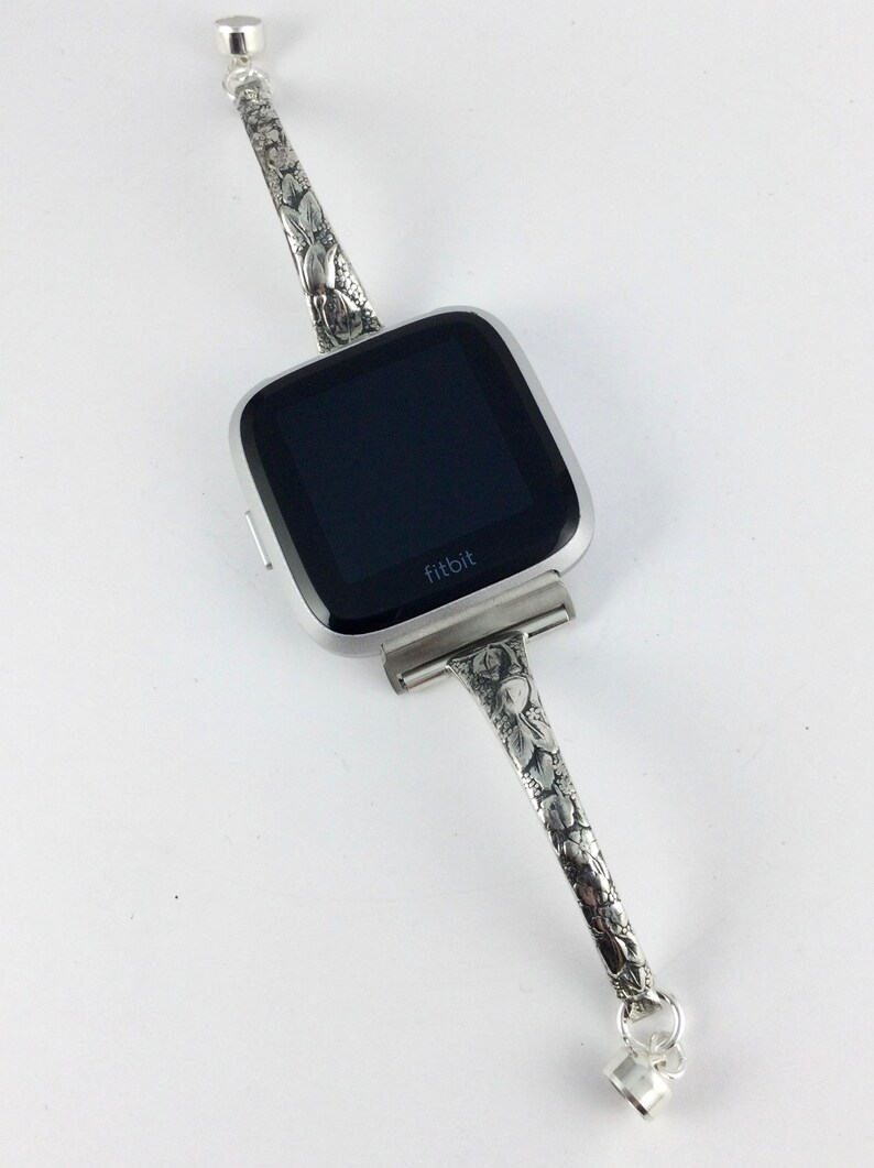May include: A silver Fitbit smartwatch with a floral patterned band. The band has a decorative charm on each end.