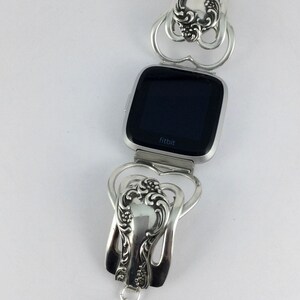 Puede incluir: Pulsera de cuchara de plata con dise&ntilde;o de coraz&oacute;n, que sostiene un reloj Fitbit plateado con una pantalla negra que dice "fitbit".