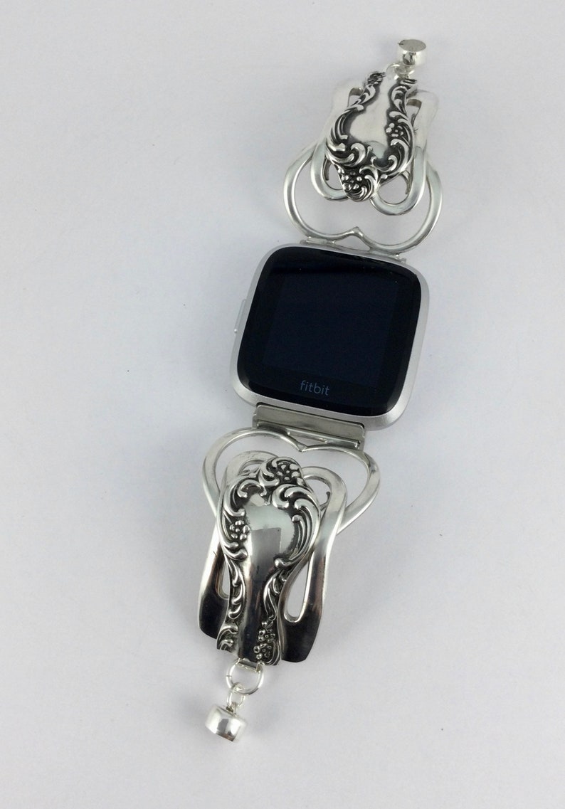 Puede incluir: Pulsera de cuchara de plata con dise&ntilde;o de coraz&oacute;n, que sostiene un reloj Fitbit plateado con una pantalla negra que dice "fitbit".