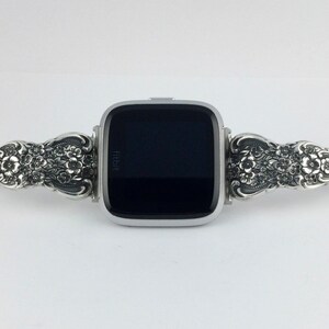 May include: Silver-toned Fitbit Versa smartwatch with a decorative floral band. The band features intricate floral and leaf patterns. The watch face is black, and the band connects with a clasp. The watch is displayed on a white background.